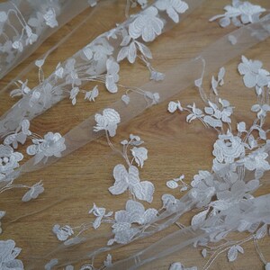May include: White floral lace fabric with detailed embroidered flowers and leaves. The delicate lace is arranged on a light background, showcasing the intricate design. Ideal for bridal wear, dresses, or decorative projects.