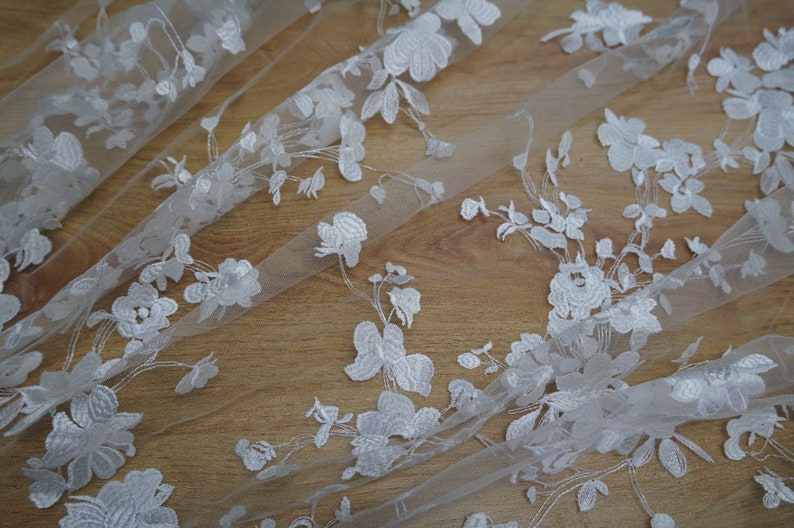 May include: White floral lace fabric with detailed embroidered flowers and leaves. The delicate lace is arranged on a light background, showcasing the intricate design. Ideal for bridal wear, dresses, or decorative projects.
