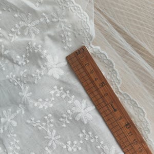 Puede incluir: Una tela blanca con bordados florales detallados y un borde festoneado, colocada junto a una regla de madera que mide 15 cm de largo. El delicado tejido muestra un patr&oacute;n repetitivo de peque&ntilde;as flores y enredaderas en toda su superficie.