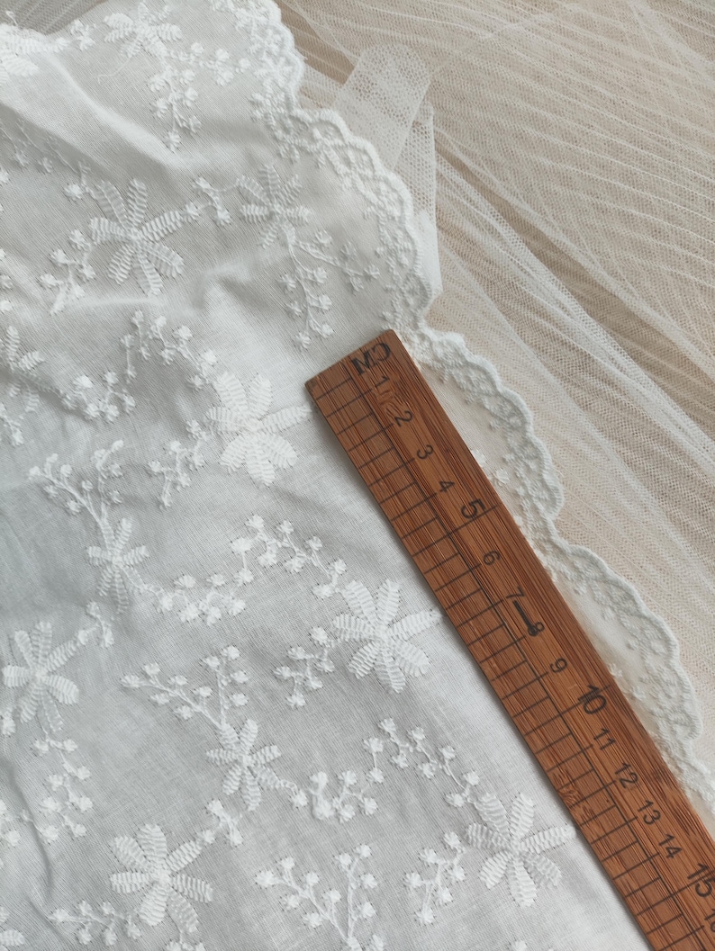 Puede incluir: Una tela blanca con bordados florales detallados y un borde festoneado, colocada junto a una regla de madera que mide 15 cm de largo. El delicado tejido muestra un patr&oacute;n repetitivo de peque&ntilde;as flores y enredaderas en toda su superficie.