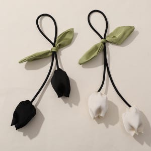 May include: Two hair ties with a floral design. Each tie features a black elastic band, a green leaf accent, and a fabric tulip in either black or white. The background is a neutral beige.