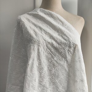 Puede incluir: Una tela blanca drapeada sobre un maniqu&iacute;, con un intrincado patr&oacute;n floral bordado en toda la superficie. El tejido muestra una textura delicada y elevada con peque&ntilde;as flores y ramitas cosidas por toda la superficie del material.