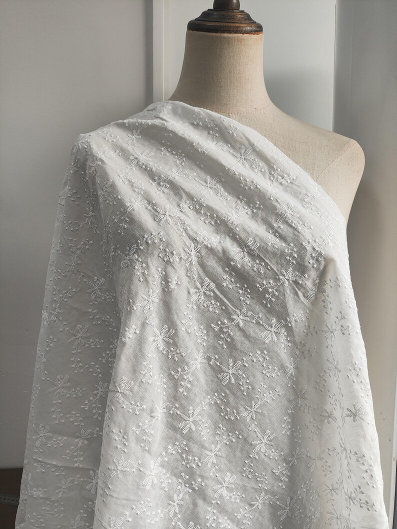 Puede incluir: Una tela blanca drapeada sobre un maniqu&iacute;, con un intrincado patr&oacute;n floral bordado en toda la superficie. El tejido muestra una textura delicada y elevada con peque&ntilde;as flores y ramitas cosidas por toda la superficie del material.