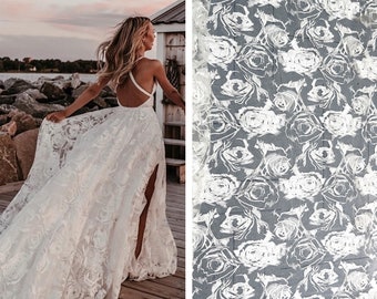 May include: A white wedding dress with a lace overlay featuring a floral pattern. The dress has a low back and a slit in the skirt. The lace pattern is also shown in a close-up.