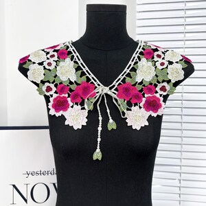 May include: A white lace collar with pink and green floral embroidery. The collar has a tie at the front.