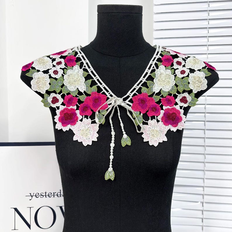 May include: A white lace collar with pink and green floral embroidery. The collar has a tie at the front.