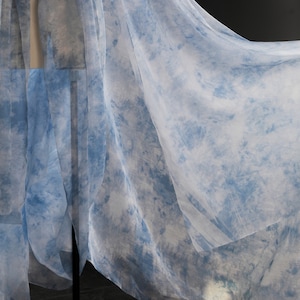 Puede incluir: Una tela transparente de color azul claro con un patrón de tie-dye. La tela está drapeada sobre una superficie y tiene una textura suave y fluida.