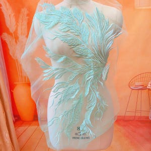 May include: A light blue lace applique with a floral design. The applique is on a white mesh background. The applique is approximately 20 cm in diameter.