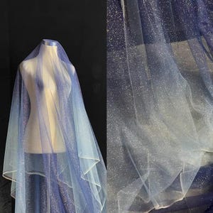 Könnte beinhalten: Ein schimmernder Stoff in Blau- und Weißtönen, drapiert über einer Schaufensterpuppe. Der Stoff hat einen Farbverlauf, der von einem tiefen Blau zu einem helleren, fast weißen Farbton übergeht. Das Material ist mit Glitzer verziert.