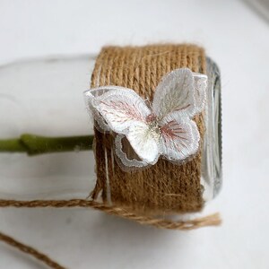 May include: A glass jar wrapped in brown twine with a white lace butterfly embellishment.