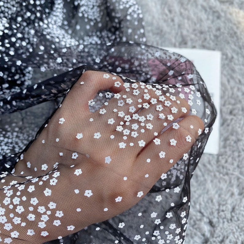 以下が含まれることがあります： 白い花柄の黒い透ける生地。生地は繊細で、柔らかく、流れるような質感です。