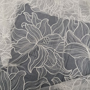 May include: White floral lace fabric with a delicate, intricate design. The lace features a repeating pattern of stylized flowers with curved lines and a textured surface.