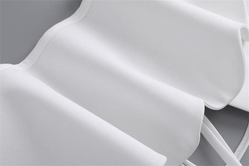 K&ouml;nnte beinhalten: Nahaufnahme eines wei&szlig;en Stoffes mit R&uuml;schendesign. Der Stoff ist gefaltet und drapiert, wodurch weiche Schatten und Glanzlichter entstehen. Die Textur erscheint glatt und die Gesamt&auml;sthetik ist sauber und elegant. Das Bild deutet auf ein Kleidungsst&uuml;ck oder Textil hin.