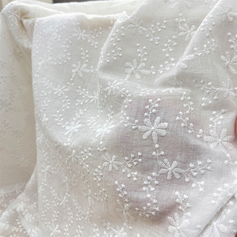Puede incluir: Una tela blanca transparente que presenta un intrincado patr&oacute;n bordado de peque&ntilde;as flores y delicados tallos similares a enredaderas. El material ligero tiene una textura suave con un motivo floral repetitivo cosido en hilo blanco.