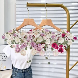 May include: A white lace collar with floral embroidery in shades of pink, purple, and green. The collar is on a hanger and has a bow tie detail.