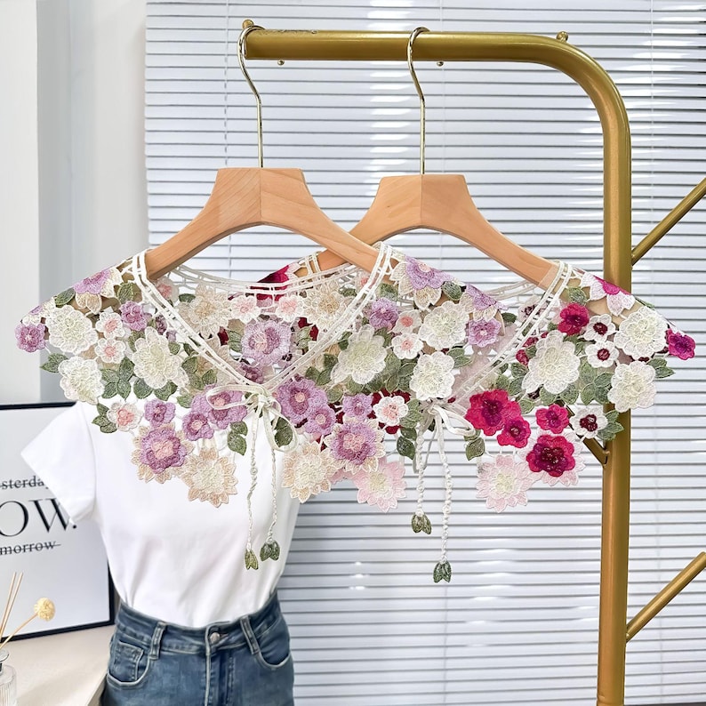 May include: A white lace collar with floral embroidery in shades of pink, purple, and green. The collar is on a hanger and has a bow tie detail.