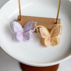 May include: Two embroidered butterfly appliques, one in a light purple and the other in a yellow color. The butterflies are on a white surface.
