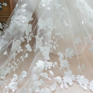 May include: White floral embroidered tulle fabric. The delicate fabric features a repeating pattern of detailed white flowers and leaves. The sheer material is draped over a mannequin, showcasing the intricate design. Ideal for bridal or formal wear.