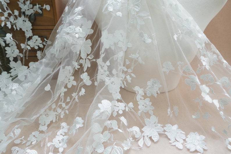 May include: White floral embroidered tulle fabric. The delicate fabric features a repeating pattern of detailed white flowers and leaves. The sheer material is draped over a mannequin, showcasing the intricate design. Ideal for bridal or formal wear.