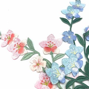 Puede incluir: Aplicación floral bordada en tonos rosa, azul y blanco con hojas verdes. Las flores tienen costuras detalladas y están dispuestas en un patrón fluido. La aplicación es probablemente para embellecer telas o proyectos de artesanía.