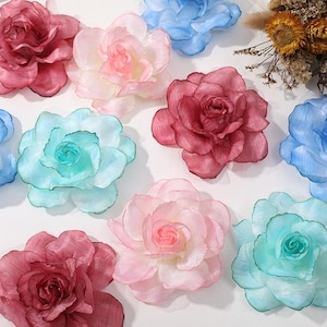 Könnte beinhalten: Eine Auswahl an farbenfrohen, durchscheinenden Stoffblumen in Rot-, Rosa-, Blau- und Türkistönen. Die Blumen sind auf einer weißen Oberfläche angeordnet, mit ein paar getrockneten Blumen im Hintergrund.