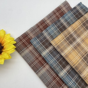 Op de afbeelding: Vier geruite stoffen stalen in tinten bruin, blauw en beige. De stoffen hebben een klassiek ruitpatroon met dunne witte lijnen. Een gele zonnebloem staat in de hoek van de afbeelding.