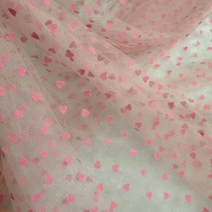 May include: Pale pink tulle fabric featuring a pattern of small pink hearts. The sheer material is semi-transparent, offering a gentle, romantic look. Suitable for various craft uses, the fabric has a delicate texture and a flowing drape.