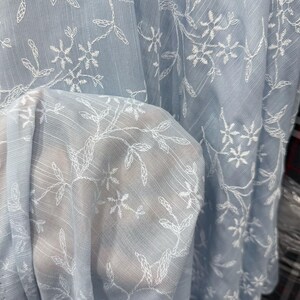 May include: A light blue sheer fabric featuring a delicate white embroidered floral pattern with thin stems and small leaves. The material has a subtle textured weave and a translucent quality that allows light to pass through the fabric layers.