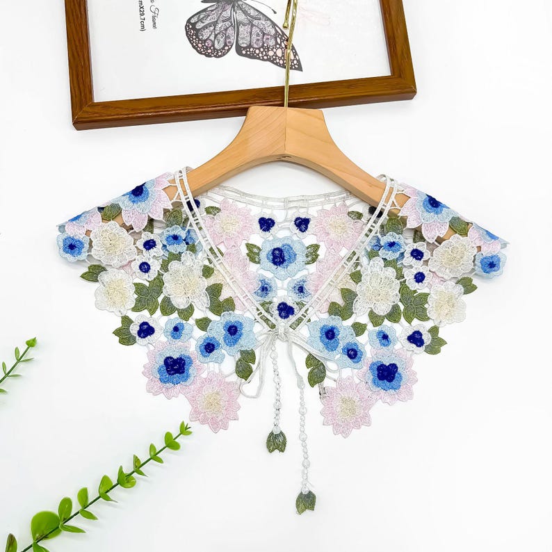 May include: A white lace collar with a floral design featuring pink, blue, and green flowers. The collar has a tie at the centre.