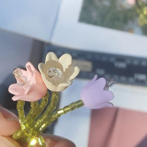 May include: A bouquet of three small flowers with green beaded stems. The flowers are made of fabric and are in shades of pink, white, and purple.