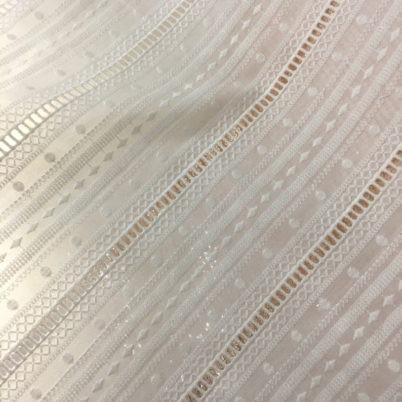 May include: White fabric with a delicate, embroidered pattern featuring a repeating geometric design. The fabric has a subtle sheen and a slightly textured surface.