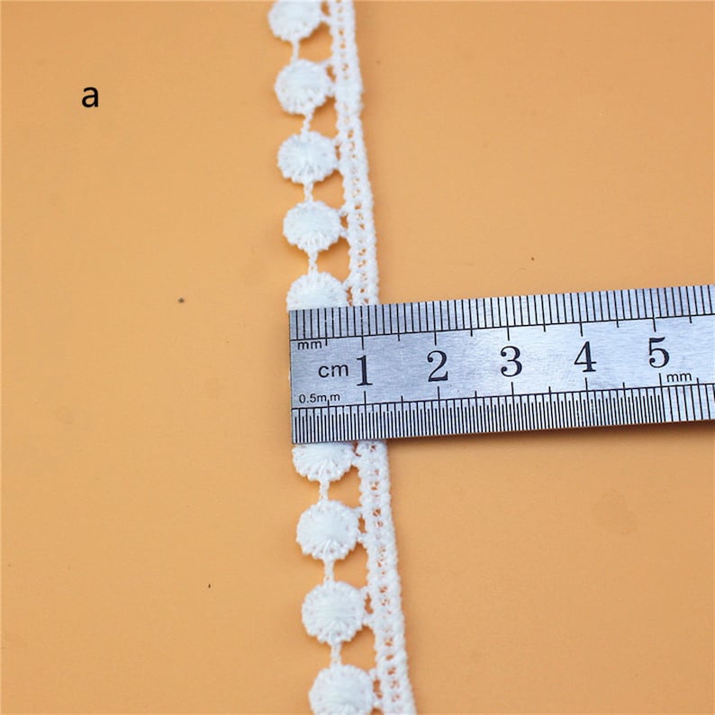May include: White cotton lace trim with a scalloped edge and small round embellishments. The trim is measured against a ruler, showing it is approximately 1 cm wide.