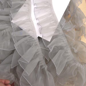 Könnte beinhalten: Nahaufnahme einer hellgrauen Rüschenborte. Die Borte besteht aus einem transparenten Stoff mit einem zarten, wellenförmigen Rand. Die Rüschen sind geschichtet und erzeugen einen voluminösen Effekt. Die Borte wird wahrscheinlich zum Nähen, Basteln oder zum Hinzufügen einer dekorativen Note verwendet.