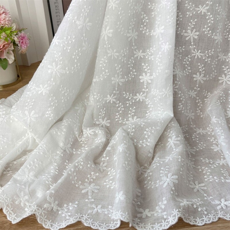 Puede incluir: Una tela blanca que presenta un intrincado patr&oacute;n de bordado floral en toda la superficie con delicadas enredaderas y flores cosidas. El material tiene una calidad semitransparente y un borde inferior festoneado con un dise&ntilde;o detallado.