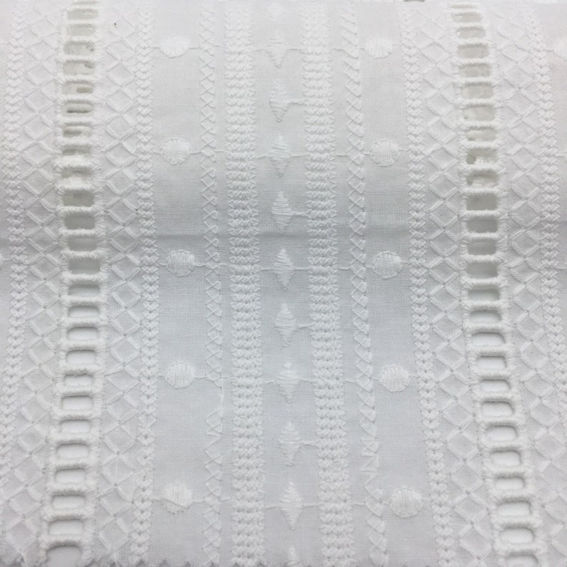 May include: White cotton fabric with an intricate embroidered pattern. The design features repeating geometric shapes and small circles.