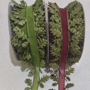 May include: Two spools of decorative ribbon featuring a central solid band flanked by intricate, three-dimensional green leaf and acorn trim. One spool has a bright lime green center, while the other features a deep burgundy red center band.