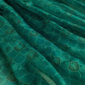 May include: A close-up of a teal fabric with a repeating fan-shaped pattern in varying shades of green and teal. The fabric is draped, creating folds and shadows that highlight the design. The overall effect is a textured, decorative textile.