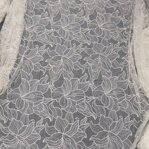 May include: White floral lace fabric with a delicate pattern of stylized flowers. The lace is sheer and has a soft, flowing texture.