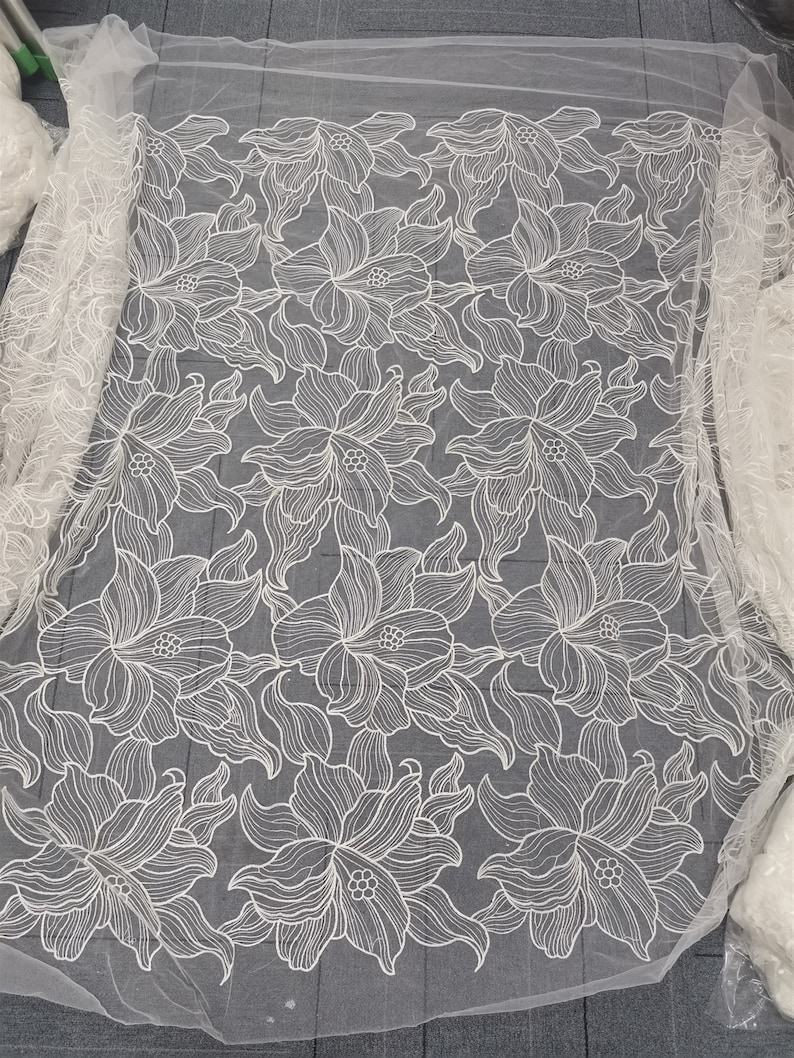 May include: White floral lace fabric with a delicate pattern of stylized flowers. The lace is sheer and has a soft, flowing texture.