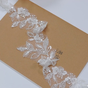 Peut inclure: Garniture en dentelle blanche avec des motifs floraux et des feuilles rehaussés de paillettes. La garniture est posée sur un fond de papier brun.