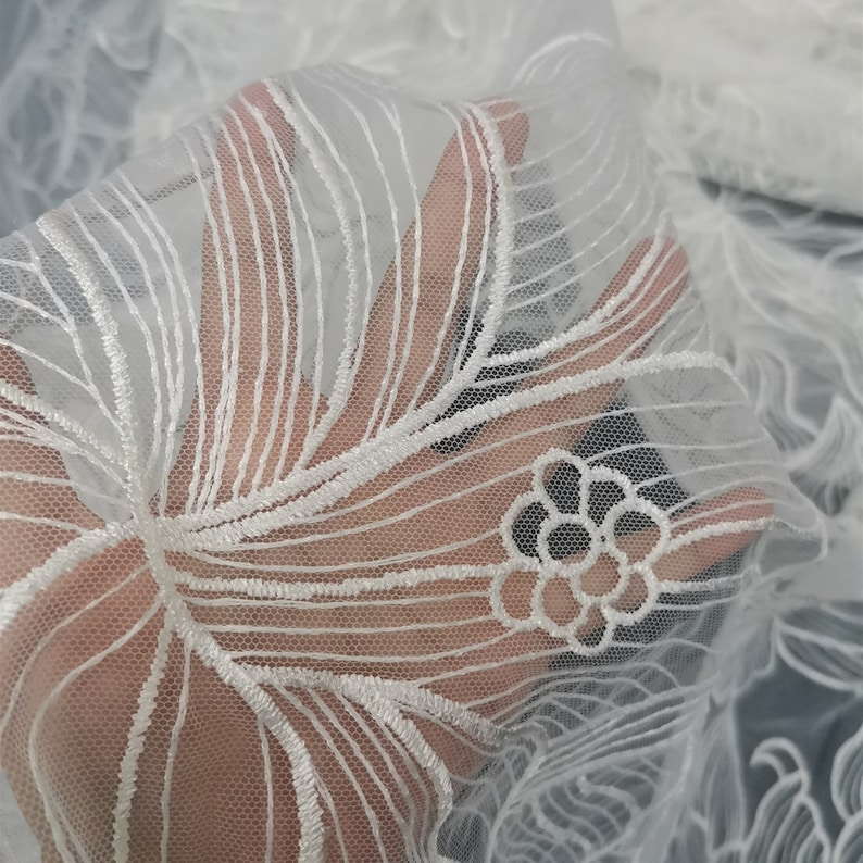 May include: White embroidered lace fabric with a floral design. The lace is delicate and intricate, with a fine mesh background.