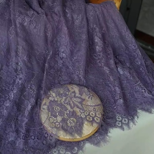Può includere: Tessuto di pizzo viola con un delicato motivo floreale. Il tessuto ha una texture morbida e fluida e una leggera lucentezza.