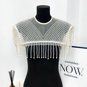 May include: A sheer, white beaded collar with fringe detail. The collar is displayed on a black mannequin, showcasing the intricate beadwork and delicate design. The collar is a fashion accessory.