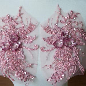 May include: Two pink floral lace appliques with beaded details. The appliques are on a sheer mesh fabric and feature delicate floral designs with pink sequins and beads.