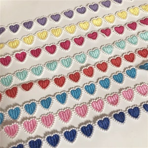 Puede incluir: Primer plano de un ribete de encaje blanco con filas de bordados en forma de corazón de colores. Los corazones son en tonos de morado, amarillo, rojo, azul y rosa.