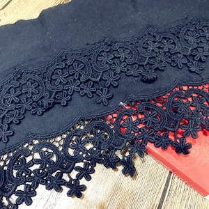 May include: Black lace trim with a scalloped edge. The lace has a floral pattern and is layered on top of a black fabric.