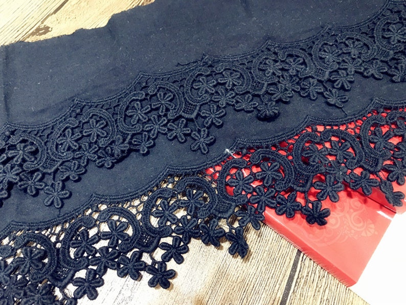 May include: Black lace trim with a scalloped edge. The lace has a floral pattern and is layered on top of a black fabric.