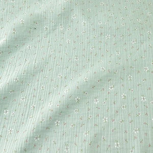 May include: A close-up of a light green fabric with a delicate floral pattern. The fabric has a crinkled texture and is covered in small white flowers with pink centers and green stems, creating a soft, textured appearance.