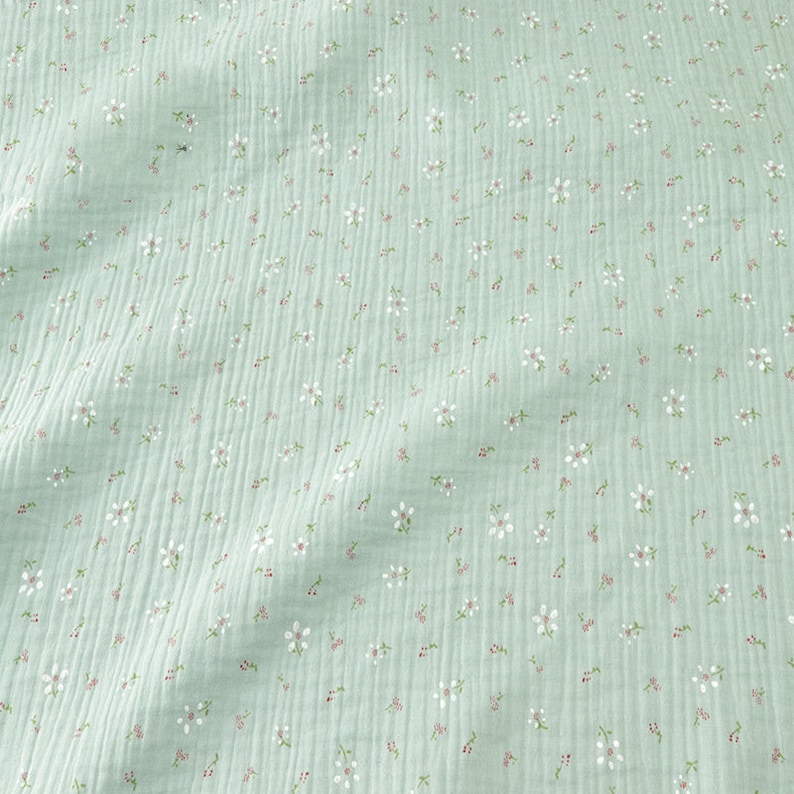 May include: A close-up of a light green fabric with a delicate floral pattern. The fabric has a crinkled texture and is covered in small white flowers with pink centers and green stems, creating a soft, textured appearance.