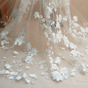 May include: Delicate white floral lace fabric. The lace features detailed, three-dimensional embroidered flowers and leaves. The fabric is semi-sheer, with a soft, flowing drape. Ideal for bridal wear, dresses, or decorative projects.